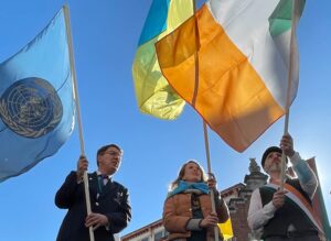 The image features a group of people gathered in a public square, holding up two large flags