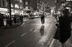 The image depicts a busy city street at night, with a man riding a bicycle down the middle of the ro