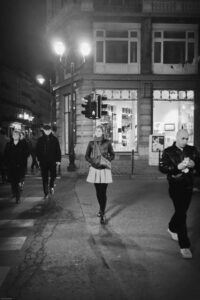 The image is a black and white photo of a group of people walking down a street at night