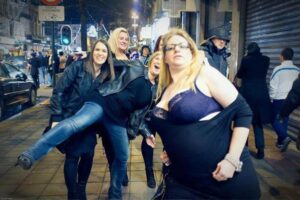 The image depicts a group of women standing on a sidewalk, posing for a picture